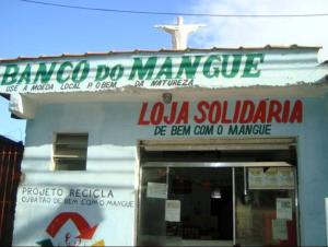 Fachada do Banco do Mangue em 2012, quando os líderes comunitários da Vila Esperança, dentre eles o Zumbi, criaram sua própria moeda comunitária: o mangue. As notas, ilustradas com animais do manguezal, circulavam entre moradores e pequenos comércios, fortalecendo a economia local. O banco nasceu para transformar lixo reciclável em valor, apoiar iniciativas sociais e incentivar que a riqueza ficasse no próprio bairro, um gesto simples que movimentou esperança, dignidade e futuro.