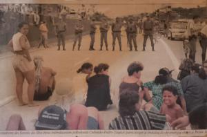 A fotografia registra a paralisação da Rodovia Padre Manoel da Nóbrega, na época chamada de Pedro Taques, organizada por lideranças comunitárias, entre elas Zumbi, como forma de reivindicar a instalação de energia elétrica na Vila Esperança. Moradores sentam-se na via principal enquanto enfrentam a presença da segurança pública. O ato evidencia a força coletiva da comunidade em defesa de condições dignas de vida.