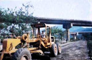 A fotografia apresenta o processo de nivelamento da Avenida Principal durante o início de sua pavimentação. Um trator trabalha na planificação do solo, enquanto a Rodovia dos Imigrantes aparece ao fundo. O registro marca um momento importante na melhoria da infraestrutura local, resultado de longa mobilização comunitária.