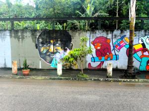 A imagem mostra o grafite em homenagem a Zumbi na fachada da Associação De Bem com o Mangue e “Asmaras” (Projeto da Gerando Falcões que visa empoderar e automatizar financeiramente as mulheres de baixa renda na comunidade), localizada na Avenida Principal da Vila Esperança. O mural retrata Zumbi segurando o mangue, símbolo da moeda que criou para incentivar tanto a preservação ambiental quanto a economia local. A obra celebra seu legado e reforça sua importância como liderança comunitária.