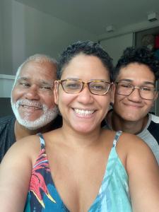 Milca entre seu pai, Antônio, e seu irmão mais novo, Henrique, durante uma visita deles à Curitiba, em 2021. Ela conta que a fotografia simboliza a conexão entre eles, na qual seu pai lhe traz muito aconchego e leveza e o irmão, apesar de não terem sido criados juntos, sempre está próximo, a apoiando e vice-versa.