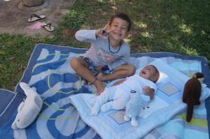 Eduardo e Gustavo, filhos de Malu, netos de João, aproveitam um momento de tranquilidade na fazenda. Gustavo está deitado sobre um pano estendido na grama, enquanto Eduardo relaxa ao seu lado.