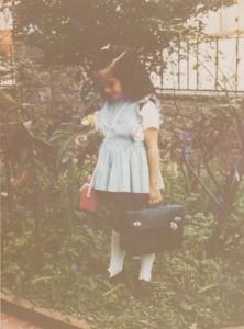 Com mais ou menos cinco anos, mais uma foto de Paula no jardim da casa, no Tucuruvi (SP). Estava indo pra escola (carregando lancheira e mala da escola), olhando para uma rosa. Como seus irmãos são mais velhos que ela, sempre brincava sozinha. Dava aula para suas bonecas, então gostava desde cedo de explicar, orientar! As brincadeiras já eram bem de organizar e ensinar.