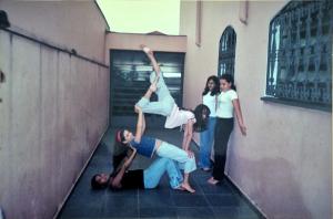 A fotografia registra um momento de liberdade e experimentação vivido no aniversário de 14 anos de Milena, no quintal de sua casa, na zona sul. Sem a presença direta de adultos, um grupo de meninas, amigas da escola e do teatro, entre risos, música e tentativas, elas encenam movimentos inspirados na ginástica artística, aprendidos com Natália, amiga e referência naquele instante, que praticava ginástica olímpica. O corpo coletivo se organiza em camadas de apoio e confiança: Milena no chão sustenta Natália, enquanto Jéssica lhe dá base. Outras duas meninas, a outra Jéssica e a Carol, permanecem observando, sem saber exatamente como participar. O gesto é lúdico, colaborativo e inventivo, mais próximo do teatro do que do esporte formal. A imagem revela uma experiência profundamente feminina, marcada pela ausência de vigilância, pela autonomia concedida pela mãe e pela ocupação livre do quintal como espaço de criação. O ato de “apresentar”, mesmo sem público definido, transforma o cotidiano em performance. A fotografia é feita pela mãe, que, ao observar a cena, decide preservá-la, reconhecendo a potência daquele instante de amizade e descoberta do próprio corpo.