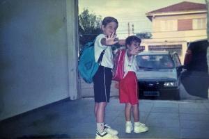 A fotografia registra o primeiro dia de Milena na primeira série, diante da escola estadual do bairro, a “escola grande”, já conhecida por meio do irmão mais velho. A imagem guarda a tensão silenciosa de um começo: o medo do desconhecido. Naquela manhã, ela sai cedo de casa ao lado do irmão Fábio. Excepcionalmente, o pai oferece carona, quebrando a rotina das longas caminhadas de cerca de vinte minutos, comuns em um bairro sem transporte público. Mais do que marcar a entrada na vida escolar, a fotografia preserva a paisagem da rua central, ponto de passagem e de encontros. É ali que a infância se estende para além dos limites da casa. A rua funciona como eixo do bairro, lugar onde todos acabam se cruzando, e sua presença na imagem amplia o valor documental da cena. O gesto capturado é simples e cuidadosamente conduzido pela mãe, que fotografa e orienta: um “tchau” sincronizado, ensaiado, quase coreografado. A memória evoca o gosto inicial pelos estudos, o vínculo afetivo com a primeira professora, Alessandra, lembrada como a mais querida, e o percurso escolar que, mais tarde, encontraria no teatro um espaço de liberdade e invenção.