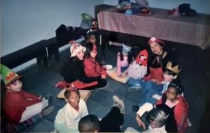 A fotografia registra um momento vivido no Neuchatel ou Centro Espírita Casa de Fabiano, no Jardim Veleiros, na Zona Sul, espaço que teve papel central na infância da Milena. O centro espírita funcionava como lugar de convivência, aprendizado e cuidado coletivo. Ali, as crianças permaneciam enquanto as mães participavam de palestras e sessões, ocupando um espaço destinado aos menores. Desde muito cedo, Milena passa a dividir a responsabilidade pelas crianças pequenas, inventando histórias, encenando pequenas peças de teatro e acolhendo bebês no colo. O centro também era um polo de atividades culturais, como aulas de flauta, violão, coral e teatro, além das ações sociais, com distribuição de sopa, roupas, remédios e brinquedos em datas comemorativas. A fotografia foi feita em um desses dias especiais. O contexto social do lugar atravessa a imagem de maneira silenciosa: Milena e sua família não se reconheciam plenamente nem como beneficiários, nem como doadores. Habitavam um espaço intermediário, marcado por necessidades reais, mas também por uma casa relativamente estável e bem localizada.