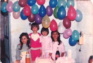 Na imagem aparecem Sandra, Juliana, Sabrina e Fabiana, em um dia marcante da infância: o aniversário de seis anos da Juliana. Foi nessa ocasião que Sandra, então com cerca de doze anos, cortou o cabelo da Juliana e da Sabrina em uma franjinha “tigelinha”, usando a tesoura que a mãe havia lhe entregue. A Sabrina gostou do resultado, achou o corte bonito e bem redondinho, enquanto a Juliana não ficou tão satisfeita. A comemoração aconteceu no Parque Santo Antônio, na casa do pai, ainda muito simples: ao fundo, vê-se o quarto pintado, sem reboco, com telhado de telha aparente. As festas eram modestas, com um bolo simples e alguns salgadinhos, mas carregadas de significado. Fotografias eram raras naquela época, já que a família não possuía câmera, um item caro para a realidade vivida. Não se sabe ao certo quem fez o registro, mas naquele aniversário houve algumas fotos, possivelmente feitas por algum amigo da família.