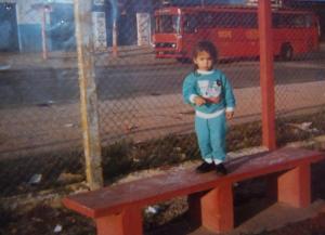 Ainda criança, Dayana aparece sobre um banco de concreto, com um ônibus vermelho da CMTC ao fundo. A imagem revela a vivência urbana desde cedo e os passeios pela cidade ainda pequena sob o olhar atento dos pais.