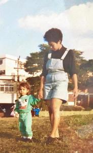 Dayana caminha de mãos dadas com sua mãe, Juju, em uma praça da cidade. A imagem revela cuidado, proteção e os primeiros passos guiados pelo afeto materno.