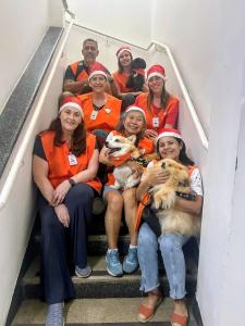 Toddy posa na escada do Hospital Mandaqui com as meninas voluntárias da ONG Terapia Cão Carinho durante a visita de Natal. A imagem revela um momento de cuidado e ternura, onde o cão voluntário e os jovens reforçam a alegria e o aconchego que podem levar às crianças hospitalizadas, tornando a data ainda mais especial.