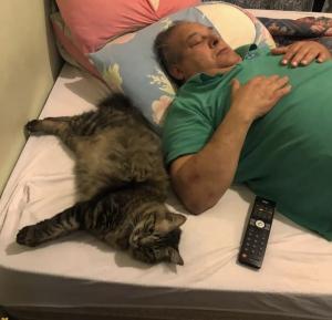 Mia, uma das gatas de Daniel, está deitada junto a José Roberto Garcia, seu pai, que já faleceu. Os dois costumavam se acomodar dessa mesma forma, dividindo o espaço e o silêncio. A imagem registra um gesto simples e recorrente, em que o corpo humano se torna apoio e companhia para o animal. Mais do que um momento de descanso, a fotografia preserva uma memória de convivência cotidiana, na qual o afeto se manifestava sem palavras, deixando vestígios de presença mesmo após a ausência.