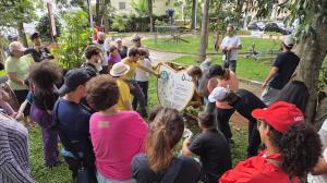 Alunos do SESC reunidos ao redor de um grande painel no Parque da Joia, acompanhando a explicação sobre o funcionamento do jogo de percurso criado para o espaço.