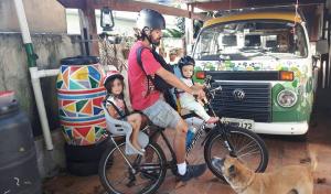 Registro de um momento feliz de Rosa e Joana durante um passeio de bicicleta com o pai, a caminho da escola. Frida, sempre presente, acompanha a família. Ao fundo, vê-se a Kombi Zeolina, companheira de inúmeras viagens de trabalho e de lazer que a família realizou por todo o Brasil.