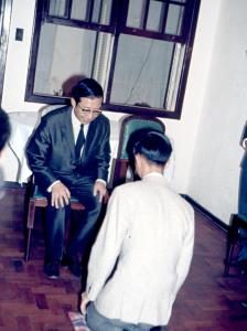 Cerimônia para ser aceito como discípulo do seu mestre, Sr. Sun, pintor de pintura chinesa clássica. Sun está sentado na cadeira. 
Cerimônia de reverência. Ele afirma que é uma cerimônia muito séria na China de antigamente. 
A cadeira vazia ao lado de Sun representa o mestre de Sun, que não pôde comparecer. 
Durante a cerimônia, deve-se ajoelhar e encostar a testa três vezes no chão, para Sun e também para a cadeira vazia.
Foi tirada na casa de Tai, com vários amigos que participaram como testemunhas.
Tai foi discípulo de 1969 a 1977, quando se mudou para Goiânia (GO).