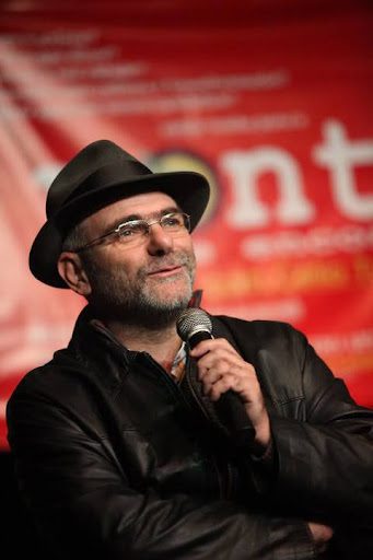 Homem de chapéu preto e óculos de grau, usando jaqueta de couro preta, segura um microfone e olha para frente. Ao fundo, um painel vermelho desfocado com texto parcialmente visível.