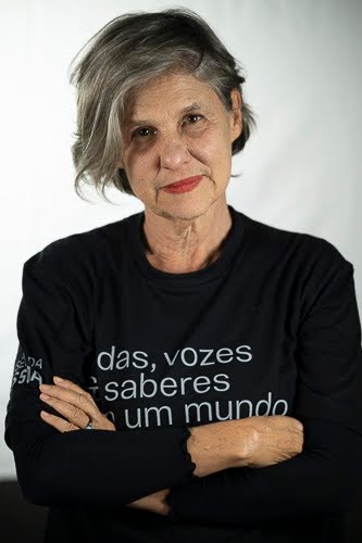 Mulher de cabelo grisalho curto e penteado de lado, vestindo camiseta preta de manga longa com texto branco estampado na frente. Ela olha diretamente para a câmera com expressão séria e braços cruzados, em frente a um fundo branco.
