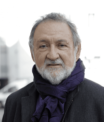 Homem branco com cabelo grisalho curto e barba branca, veste casaco escuro e cachecol roxo. Ele olha diretamente para a câmera, com expressão neutra. O fundo é desfocado, mostrando formas urbanas e luz natural.