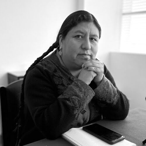 Mulher de expressão séria está sentada, apoiando o queixo nas mãos entrelaçadas. Ela tem cabelos longos presos em uma trança e usa brincos pequenos. Veste um casaco escuro com detalhes bordados. Sobre a mesa à sua frente há um caderno e um celular. A fotografia é em preto e branco e mostra um ambiente interno iluminado por uma janela ao fundo.