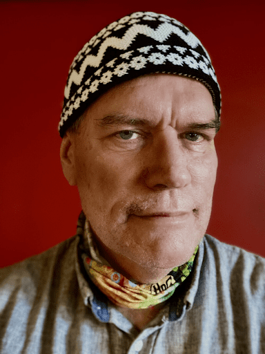 Um homem de pele clara e expressão séria olha diretamente para a câmera. Ele usa um gorro de crochê preto e branco com padrão geométrico e floral. No pescoço, traz um lenço colorido com tons de verde, amarelo e laranja, por baixo de uma camisa cinza clara de botões. O fundo é completamente vermelho.