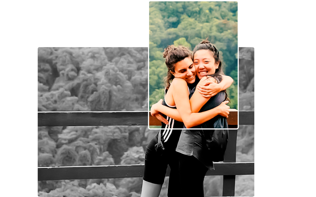 Fotografia em preto e branco. Duas mulheres se abraçam, sorrindo. A primeira, tem o cabelo castanho enrolado, preso parcialmente atrás. Ela usa short e blusa preta. A segunda, tem o cabelo preto, preso no alto da cabeça. Ela veste calça preta, blusa cinza e tem uma mochila preta nas costas. Ao fundo, há uma cerca e mata. Um retângulo transparente, com margem branca, que está preenchido cores, revela a mata ao fundo até a barriga das jovens.