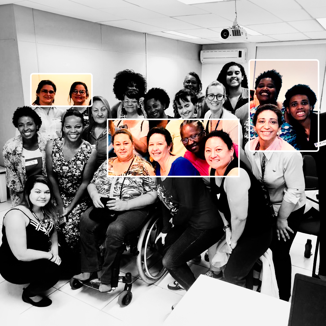 Fotografia em preto e branco. Um grupo de mulheres está em pé, olhando para frente e sorrindo. Elas estão uma ao lado da outra e ao centro, há uma mulher na cadeira de rodas. Atrás, há paredes e tetos brancos, uma câmera de segurança e um ar-condicionado. Há três quadrados transparentes, com margem branca, que está preenchido por cores que revelam cores entre algumas das mulheres.