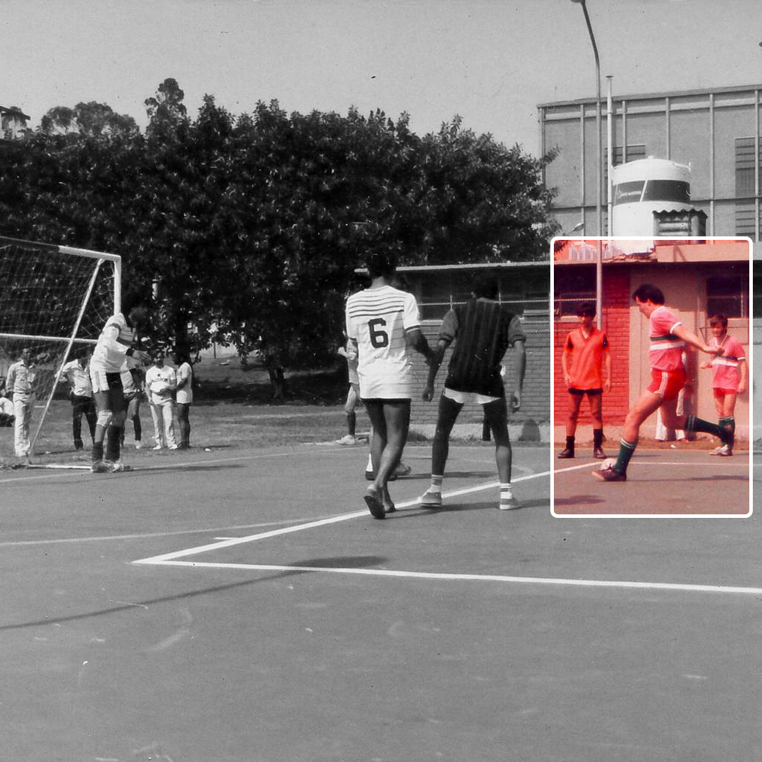 Fotografia em preto e branco. Dez homens, espalhados em um campo de futsal, se movimentam. Na esquerda, há a trave do gol. Todos usam camiseta, short, meia e tênis. Ao fundo, há algumas árvores e um prédio pequeno. Um retângulo, com margem branca, é preenchido com predominância vermelha, que transparece um homem em movimento com a bola ao lado do seu pé esquerdo, enquanto o direito e seus braços estão para trás.