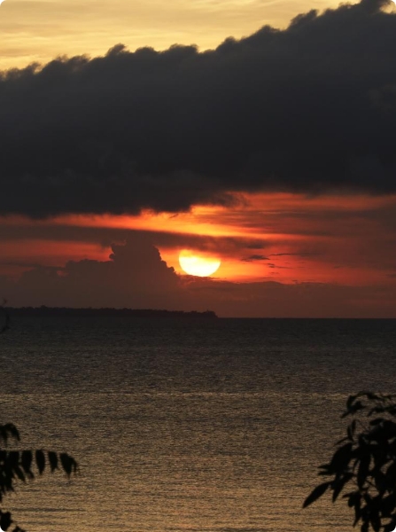 Fotografia colorida. Acima do mar, há um sol atrás de nuvens. Ao fundo, há folhagens.