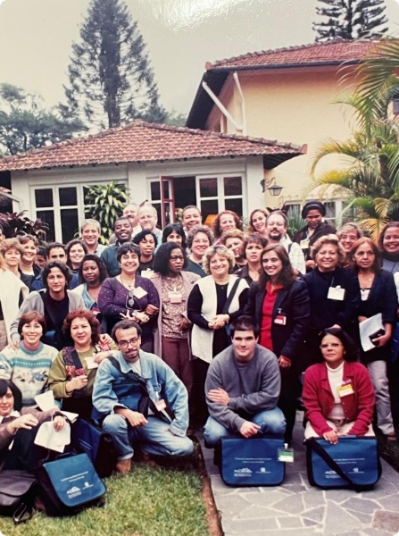 Fotografia colorida. Um grupo de pessoas se reúne em frente a uma casa branca, com duas janeiras. Atrás da casa, há árvores. Todos olham para frente, sorrindo.