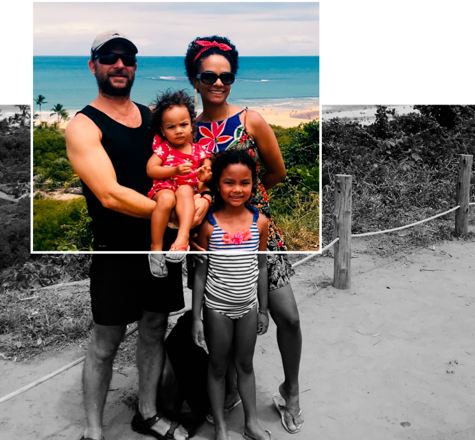 Fotografia em preto e branco. Ao fundo, há grama, areia e mar. Um homem de camiseta e bermuda preta segura uma criança no colo. Ele usa boné branco e óculos de sol preto. A criança, no colo, usa um vestido vermelho floral. Ao lado deles, uma mulher de cabelos enrolados e curto, usa um vestido azul florido, óculos de sol preto e um arco vermelho com lacinho em cima na cabeça. Na sua frente, há uma menina de maiô branco e azul listrado. Um quadrado transparente, com margem branca, que está preenchido por cores, revela as cores desde o mar ao fundo até o peito da menina de maiô.