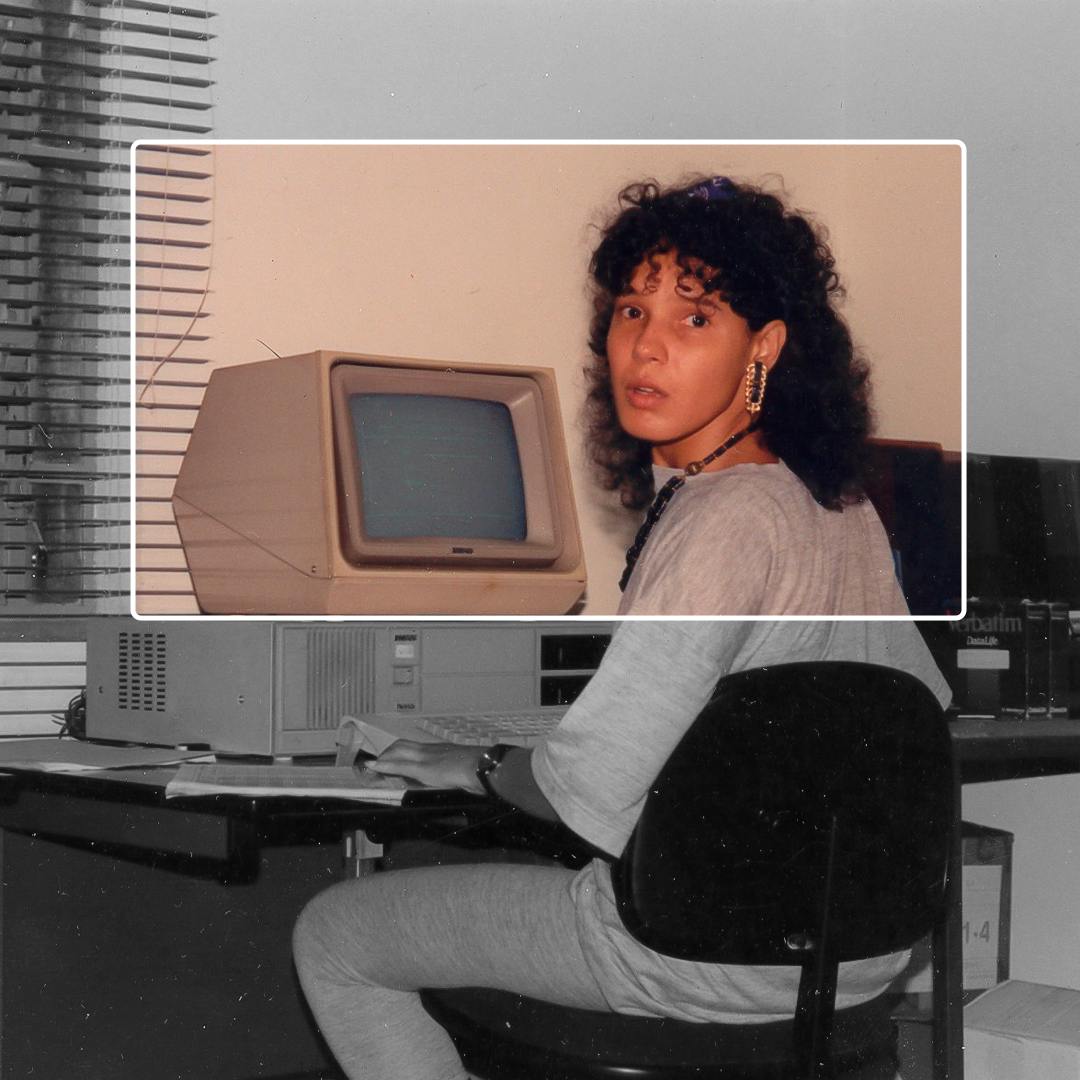 Fotografia em preto e branco. Uma moça está sentada em uma cadeira de frente para uma mesa, com um computador que está em cima de um CPU. Ela olha para trás, por cima do ombro. Tem os cabelos enrolados e pretos, longos. Usa uma camiseta e calça cinzas. Um triângulo transparente, com margem branca, que está preenchido por cores, revela de forma mais forte as cores do computador e do ombro para cima da moça.