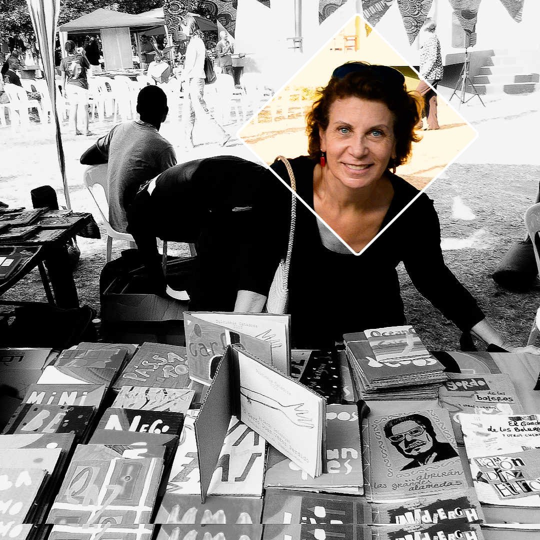 Fotografia em preto e branco. Uma mulher olha para frente e sorri. Ela está inclinada levemente para frente, onde há uma mesa cheia de livros. Ao fundo, há cadeiras e mais mesas com livros e pessoas. Ela usa uma blusa preta e tem os cabelos curtos e castanhos. Um retângulo transparente, com margem branca, que está preenchido por cores, revela de forma mais forte as cores do rosto dela.