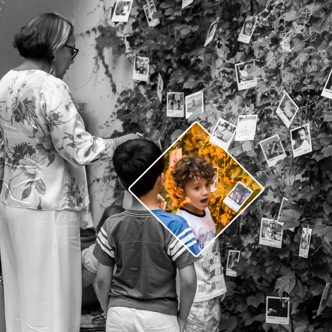 Fotografia em preto e branco. Uma senhora em pé, de costas, está com a mão direita encostado em uma parede de folhagens e fotos penduradas. Ao seu lado, há dois meninos, eles estão um de frente para o outro. O segundo menino, está de costas, o primeiro, de lado, em frente a parede, de boca aberta. Um triângulo transparente, com margem branca, que está preenchido por cores, revela de forma mais forte as cores do rosto do primeiro menino.