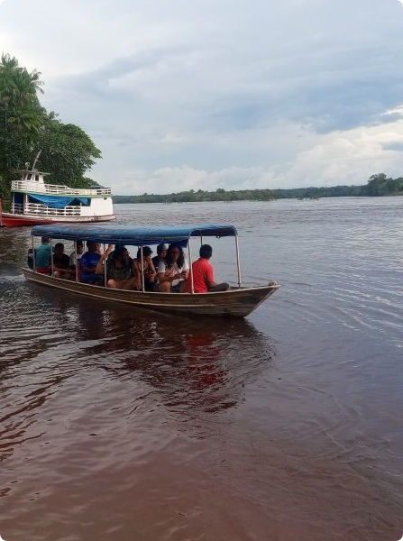 Fotografia colorida. Um barco, com pessoas dentro, avança em um rio. Ao fundo, há mata e um céu coberto por nuvens.