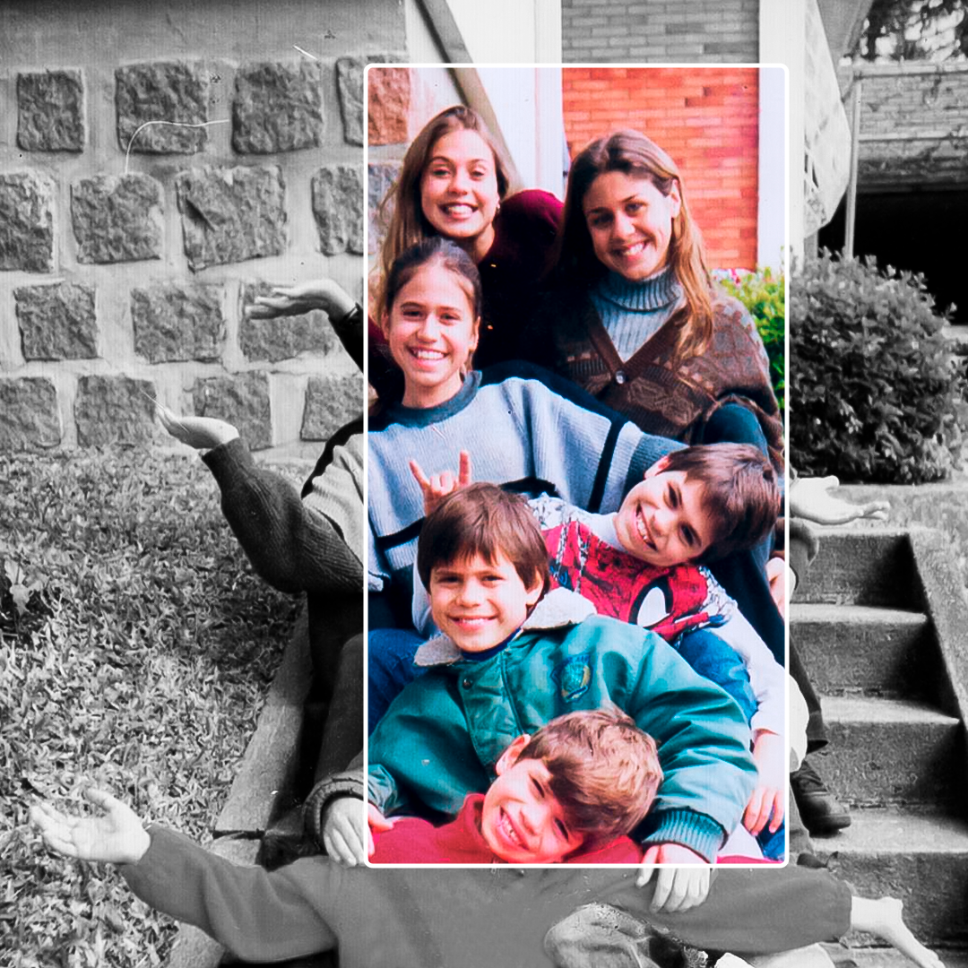 Fotografia em preto e branco. Em uma escada, há três crianças, uma em frente a outra. Atrás dos meninos, há três jovens, também uma atrás da outra, atrás deles na escada. Todos estão sorrindo e olhando para frente, com os braços esticados para o lado, com a cabeça também inclinada para o lado diferente da pessoa à frente. O primeiro menino é loiro e tem os cabelos lisos, com franjinha na testa, usa um moletom vermelho. O segundo menino tem o cabelo castanho-claro liso, franjinha na testa, usa uma jaqueta verde. O terceiro menino tem o cabelo castanho-claro liso, franjinha na testa, usa uma camiseta do homem-aranha. Atrás, a jovem tem o cabelo preso para trás e usa uma blusa longa azul-claro com listas azuis escuras. Atrás dela, há uma moça com jaqueta marrom estampada e dentro uma azul clara com gola rolê. Por última, uma menina loira usa uma camisa longa vermelha. Ao centro da fotografia, há um retângulo transparente, com margem branca, que abrange todas as pessoas e está preenchido por cores da fotografia.