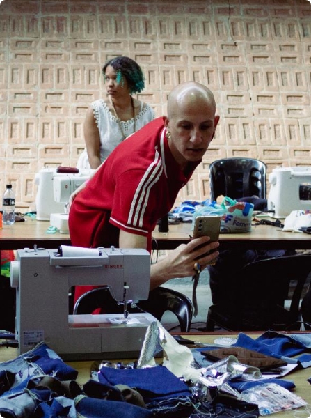 Fotografia colorida. Um homem, está inclinado para uma máquina de costura, à sua frente. A máquina está em cima de uma mesa, com tecidos azuis. O homem é careca e usa uma camiseta vermelha. Atrás, há uma mulher em pé, atrás de uma mesa com máquinas de costura.